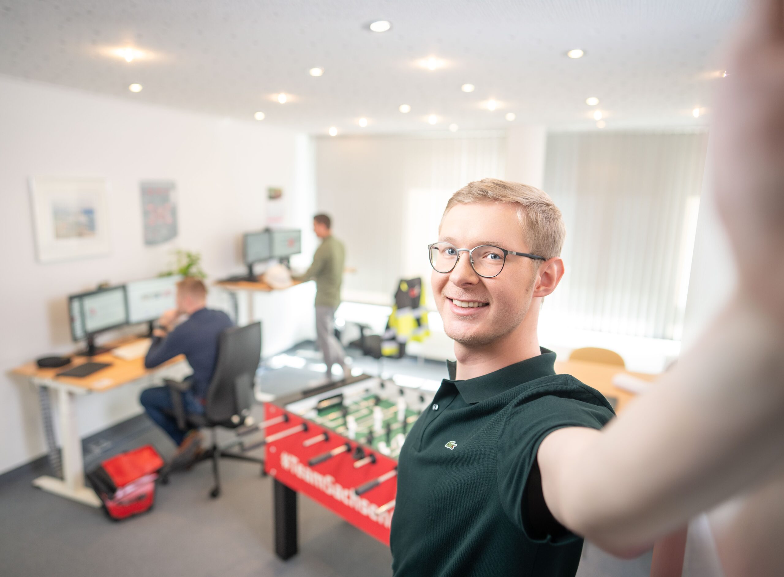 A young man takes a selfie in a modern office with colleagues and a table football game.